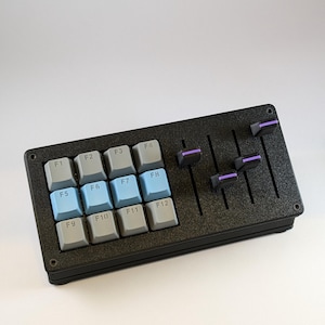 Op de afbeelding: Zwart toetsenbord met 12 functietoetsen met de labels F1 tot en met F12. De toetsen zijn gerangschikt in een 3x4 raster. Het toetsenbord heeft een getextureerd oppervlak en zes zwarte faders met paarse accenten.