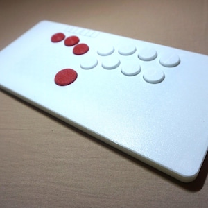 Może przedstawiać: Biały prostokątny kontroler do gier z czerwonymi i białymi przyciskami. Kontroler ma w sumie 13 przycisków.