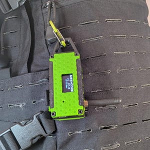 May include: A green and black electronic device with the text "MESHCORE v1.12.0" displayed on a small screen. The device is attached to a black backpack with a carabiner clip. An antenna extends from the device.