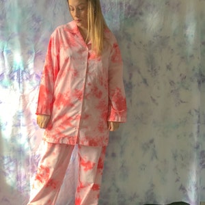 May include: Pink tie-dye pajama set. The set includes a button-down shirt with a collar and long sleeves, and matching pants. The pajamas have a light pink base with vibrant red-pink tie-dye patterns. The fabric appears soft and comfortable.