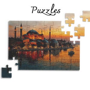 Può includere: Un puzzle che raffigura una vista al tramonto della Basilica di Santa Sofia a Istanbul, con le sue cupole e i minareti riflessi nell'acqua. La parola Puzzles è stampata in corsivo nero sopra i pezzi del puzzle parzialmente assemblati.