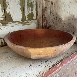 May include: A large, wooden bowl with a natural wood finish. The bowl has a slightly reddish hue and shows signs of age and use. It is sitting on a wooden shelf with a white and red paint finish.