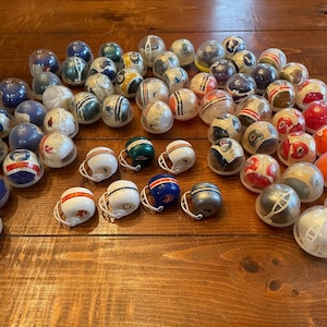 Peut inclure: Une collection de casques de football miniatures de différentes couleurs d'équipe et logos, y compris le rouge, le blanc, le bleu, le vert et l'orange. Les casques sont disposés en cercle sur une surface en bois.