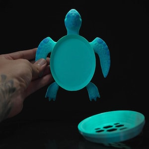Puede incluir: Un plato de cerámica de color azul turquesa en forma de tortuga con una parte superior redonda y plana y un borde elevado. La tortuga tiene un caparazón y patas detalladas.