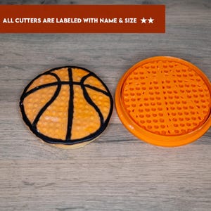 May include: Orange and black basketball cookie cutter with a textured surface. The cutter is next to a basketball cookie with orange icing and black lines.