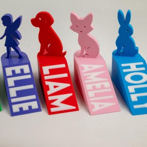 May include: Five colorful doorstops with animal and fairy figures. The doorstops are green, blue, red, pink, and blue, each with a different name in white lettering: THOMAS, ELLIE, LIAM, AMELIA, and HOLLY. The figures include a dinosaur, fairy, dog, cat, and rabbit.
