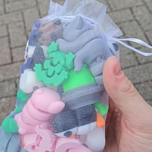 May include: A collection of colorful foam shapes, including a pink pig, a green tree, and a gray hippo, are contained within a sheer white drawstring bag. The bag is held in a person's hand, with a brick surface in the background.