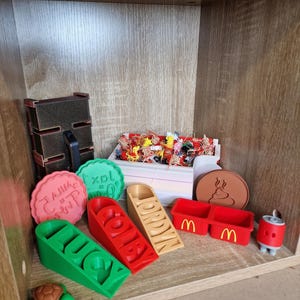 May include: A variety of 3D-printed items are displayed on a wooden shelf. Items include name blocks, a turtle figurine, a McDonald's container, and a brown coaster with a poop emoji. Other items include a red and black box and a can.