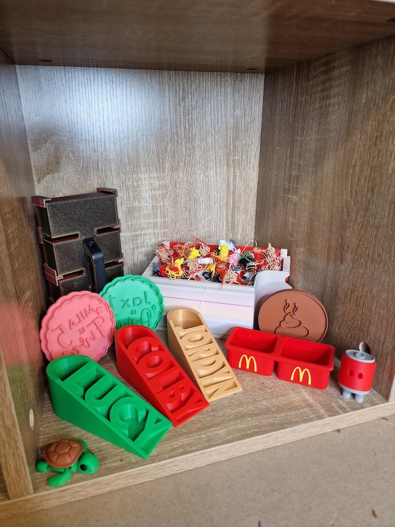May include: A variety of 3D-printed items are displayed on a wooden shelf. Items include name blocks, a turtle figurine, a McDonald's container, and a brown coaster with a poop emoji. Other items include a red and black box and a can.