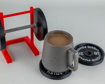 Puede incluir: Una taza de café de cerámica gris con un acabado moteado y un asa negra se asienta sobre un posavasos de placa de peso negro. La taza está llena de café marrón. Un estante de pesas rojo con una barra de pesas gris y placas de pesas negras está en el fondo.