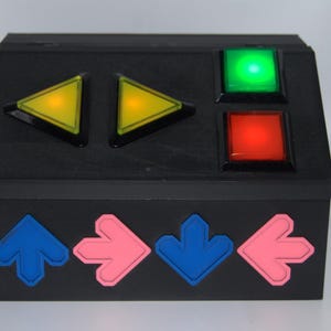 Könnte beinhalten: Eine schwarze Box mit vier Pfeiltasten unten und drei farbigen Tasten oben. Die Tasten sind gelb, grün und rot.