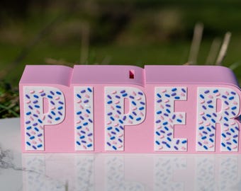 May include: Pink personalized name block with the name "PIPER" spelled out in white letters. The block is decorated with a sprinkle pattern in pink, blue, and white.