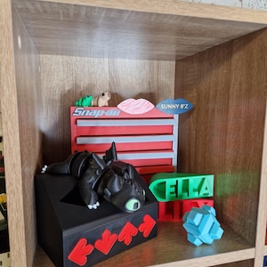 May include: A wooden shelf displays various 3D-printed objects. A black dragon figure rests on a black box with red arrow accents. Other items include a red and blue toolbox, a green nameplate that says "ELLA", and a blue geometric puzzle.