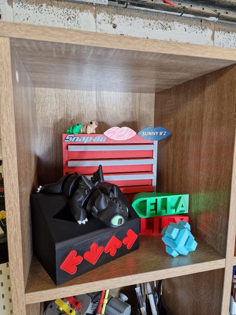 May include: A wooden shelf displays various 3D-printed objects. A black dragon figure rests on a black box with red arrow accents. Other items include a red and blue toolbox, a green nameplate that says "ELLA", and a blue geometric puzzle.