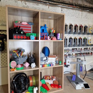 May include: A wooden shelf unit filled with various 3D-printed objects in a workshop setting. Items include a black bust, colorful spools of filament, and miniature models. The shelf also displays a 3D printer and paint supplies.