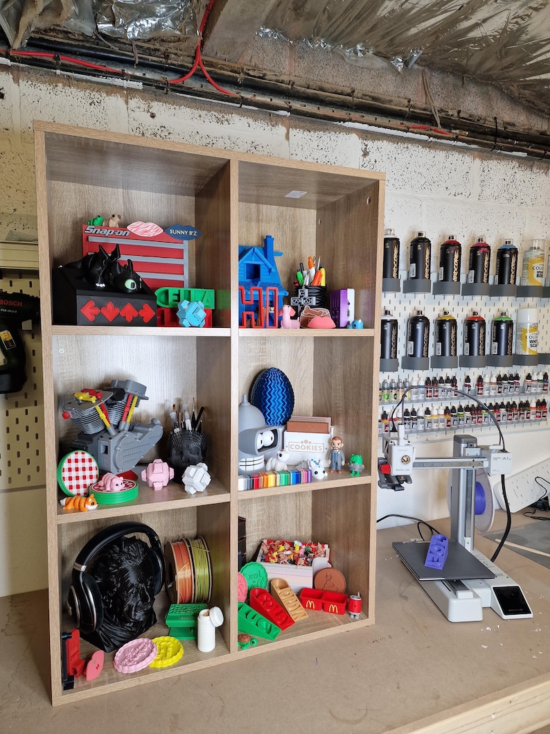 May include: A wooden shelf unit filled with various 3D-printed objects in a workshop setting. Items include a black bust, colorful spools of filament, and miniature models. The shelf also displays a 3D printer and paint supplies.