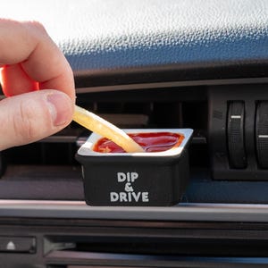 May include: A black and white car accessory with the text "Dip & Drive" on the front. The accessory is a small container for holding dip or sauce. A hand is dipping a french fry into the container.