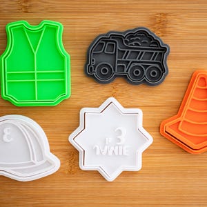 May include: Set of five cookie cutters in various shapes and colours, including a hi-vis vest, a dumper truck, a traffic cone, a hard hat, and a star-shaped cutter with the text "It's 3" in the centre.