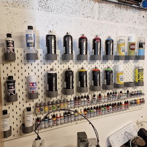 May include: A collection of spray paint cans and paint bottles organized on a white pegboard. The cans are black with various color accents and the text "ARDCORE". Bottles of paint are arranged below, with a 3D printer in the foreground.