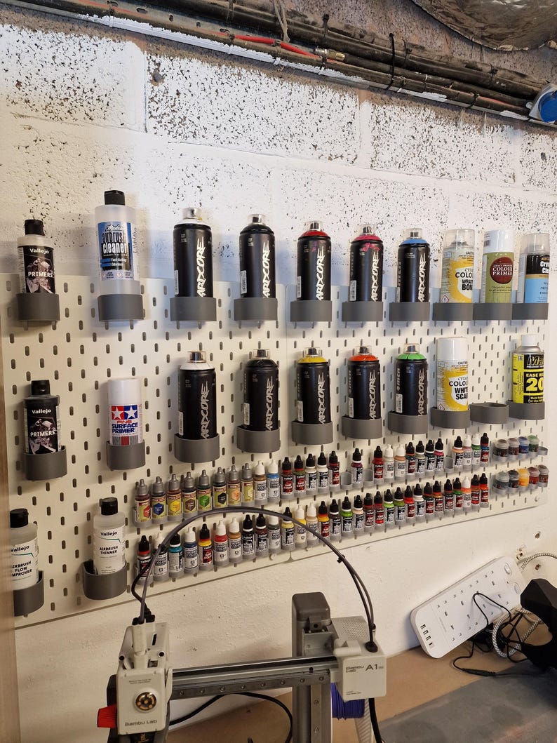 May include: A collection of spray paint cans and paint bottles organized on a white pegboard. The cans are black with various color accents and the text "ARDCORE". Bottles of paint are arranged below, with a 3D printer in the foreground.