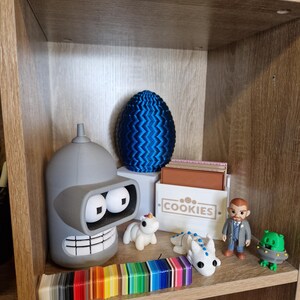 May include: A wooden shelf displays various figurines and objects. A gray Bender figurine, a blue patterned egg, and a box labeled "COOKIES" are prominent. A rainbow color swatch and several small toys are also visible, creating a diverse collection.