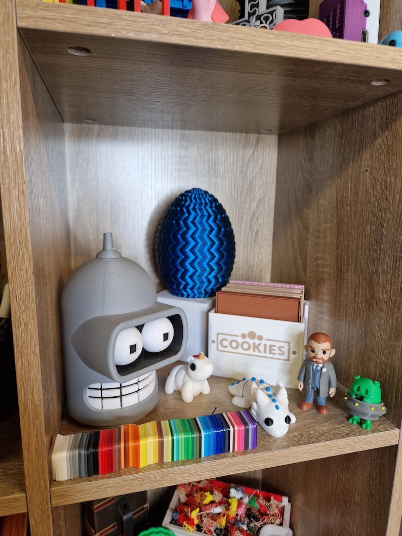 May include: A wooden shelf displays various figurines and objects. A gray Bender figurine, a blue patterned egg, and a box labeled "COOKIES" are prominent. A rainbow color swatch and several small toys are also visible, creating a diverse collection.