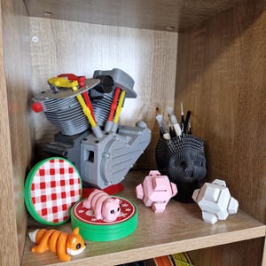 May include: A shelf displays 3D-printed items. A gray engine model, a red and white checkered coaster, a pink pig figurine, and a segmented orange fox are visible. A black skull-shaped pen holder and geometric shapes are also present.