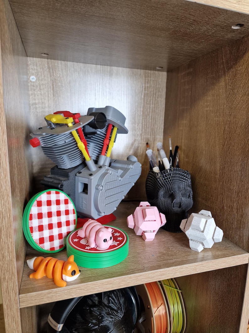 May include: A shelf displays 3D-printed items. A gray engine model, a red and white checkered coaster, a pink pig figurine, and a segmented orange fox are visible. A black skull-shaped pen holder and geometric shapes are also present.