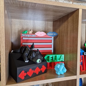 May include: A wooden shelf unit displays various 3D-printed items. A black dragon figure rests on a black box with red arrow accents. Other items include a green nameplate that says "ELLA", a blue puzzle, and a red and gray toolbox with the text "Snap-on".