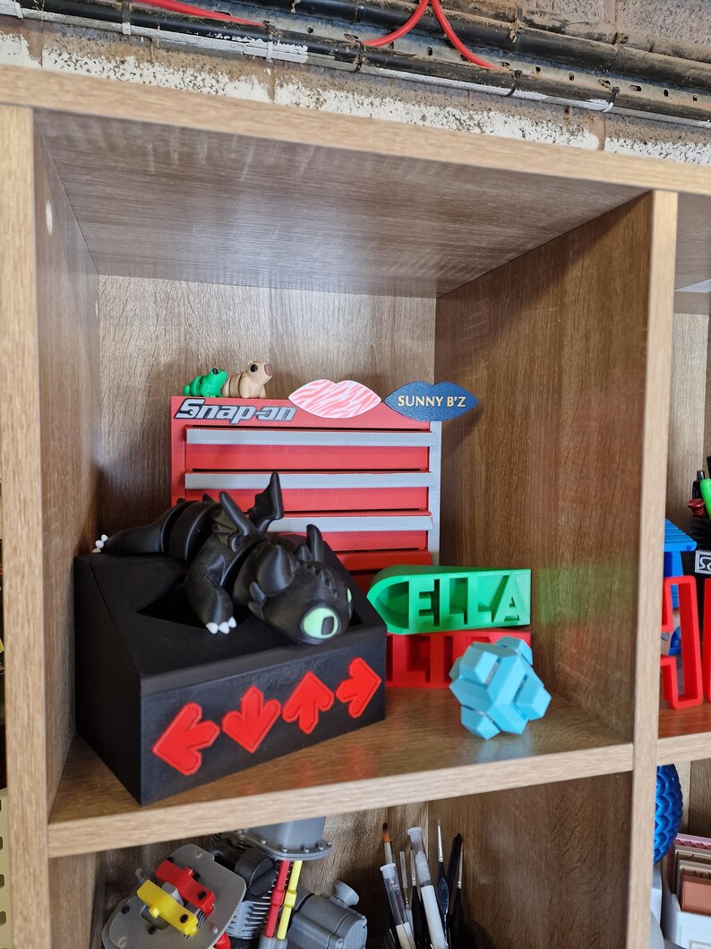 May include: A wooden shelf unit displays various 3D-printed items. A black dragon figure rests on a black box with red arrow accents. Other items include a green nameplate that says "ELLA", a blue puzzle, and a red and gray toolbox with the text "Snap-on".