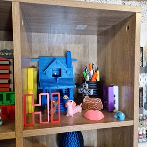 May include: A wooden shelf displays various 3D-printed items. A blue house, a red "HI" sign, and a pink dachshund are among the objects. Other items include a dog bowl, a blue sphere, and pens in a holder.
