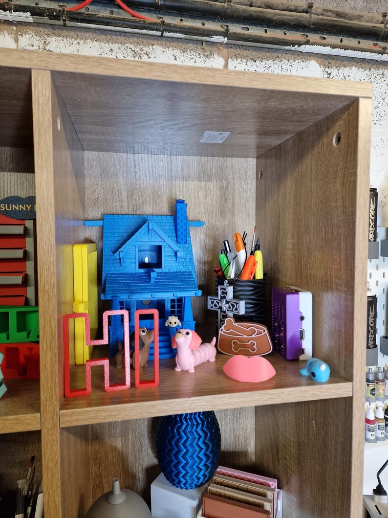 May include: A wooden shelf displays various 3D-printed items. A blue house, a red "HI" sign, and a pink dachshund are among the objects. Other items include a dog bowl, a blue sphere, and pens in a holder.