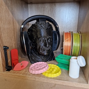 May include: A black bust of Albert Einstein wearing black headphones. The shelf also holds a variety of items, including spools of orange and green wire, pink and yellow cookie cutters, and a white container.