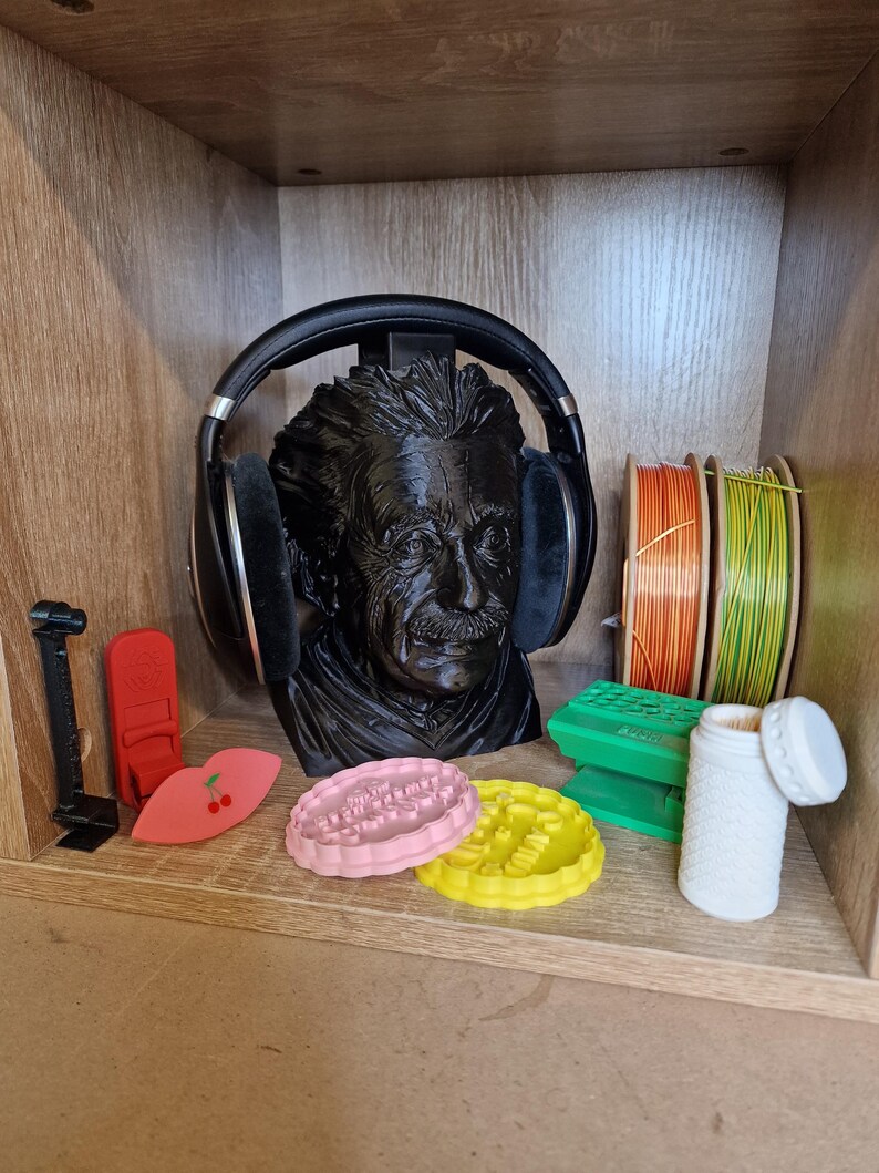 May include: A black bust of Albert Einstein wearing black headphones. The shelf also holds a variety of items, including spools of orange and green wire, pink and yellow cookie cutters, and a white container.