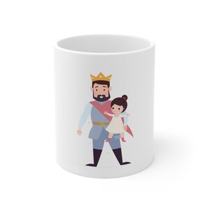May include: A white ceramic mug featuring a colorful cartoon illustration of a bearded king wearing a yellow crown, a light blue tunic, and a red cape, holding a small child with dark hair in a white dress and red boots against a plain background.