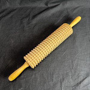 Könnte beinhalten: Eine hölzerne Teigrolle mit einer strukturierten Oberfläche aus sich wiederholenden dreieckigen Formen. Die Griffe sind glatt und abgerundet. Die Teigrolle hat eine helle, natürliche Holzfarbe.