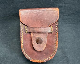 Puede incluir: Una funda de cuero marrón con cierre de solapa y botón. La funda tiene un borde cosido con hilo amarillo. El cuero parece desgastado, lo que sugiere una estética vintage. Diseñado para guardar objetos pequeños.