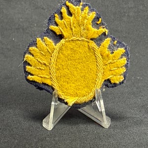 May include: A fabric patch featuring a central yellow oval surrounded by yellow embroidered wings and a flame-like crest at the top. The design is set against a dark blue background with frayed edges, showcasing detailed yellow thread embroidery.