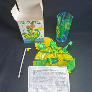 May include: A vintage "Mr. Turtle" floating glass caddy set. The set includes a box with a turtle graphic, a clear glass with turtle designs, an inflatable yellow and green turtle, a straw, and instructions.