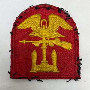 Può includere: Una toppa ricamata rossa con un emblema color oro raffigurante un'aquila appollaiata su un fucile, posizionato orizzontalmente sopra un'ancora. La toppa ha un bordo superiore arrotondato e fili neri sciolti visibili lungo il perimetro.