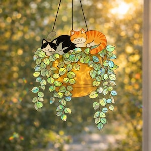 May include: A hanging stained glass art piece featuring a yellow planter with cascading green leaves. Two cats, one black and white, the other orange tabby, rest atop the planter. The background is a blurred view of sunlight and foliage.