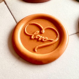 May include: An assortment of wax seals in various colors, including orange, brown, and green. Each seal features the word "love" in a cursive font within a heart shape. The seals are arranged on a light-colored surface.