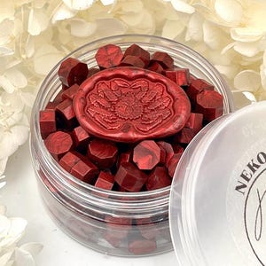 Könnte beinhalten: Ein durchsichtiges Plastikglas gefüllt mit roten Wachsperlen und einem dekorativen Wachssiegel. Das Siegel hat ein florales Design. Der Deckel des Glases ist teilweise sichtbar, mit dem Text "NEKO Studio" und einer Katzen-Silhouette.