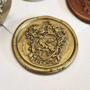 May include: A gold-colored wax seal featuring a detailed embossed crest with a bird at the center and the text RAVENCLAW inscribed on a banner at the bottom. The circular seal has a raised border and is surrounded by other wax seals in various colors.