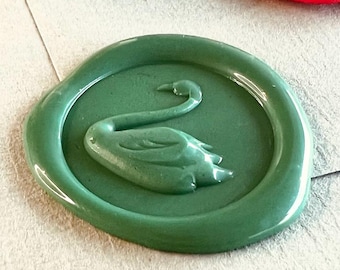 Può includere: Un timbro sigillo di cera con un disegno di cigno, insieme a sigilli di cera rossi, bianchi, verdi e bronzo. Il disegno del cigno è in rilievo sul timbro e sui sigilli di cera. Gli oggetti sono su una superficie verde chiaro.