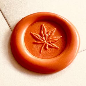 Könnte beinhalten: Eine Sammlung farbiger Wachssiegel mit einem Ahornblatt-Design. Die Siegel sind in den Farbtönen Orange, Rot, Grün und Braun gehalten. Ein goldfarbener Metallstempel mit dem gleichen Blattdesign ist ebenfalls vorhanden.