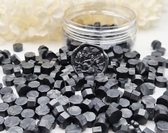 May include: A collection of small, black, hexagonal wax seal beads scattered around a clear plastic jar. One black wax seal impression featuring a detailed embossed design of a figure on horseback is placed prominently in the center of the pile.