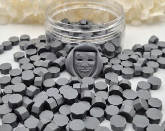 May include: A collection of small, gray, octagonal wax seal beads scattered around a clear plastic jar. One larger, gray wax seal bead features a detailed, molded Guy Fawkes mask design with a hood, resting prominently in the center of the pile.