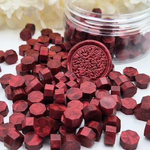 Op de afbeelding: Een verzameling kleine, achthoekige donkerrode waszegelkralen met een gemarmerde textuur, verspreid naast een doorzichtige plastic pot gevuld met kralen en een enkele ronde waszegel met een ingewikkeld, in reliëf gemaakt bloemmotief.