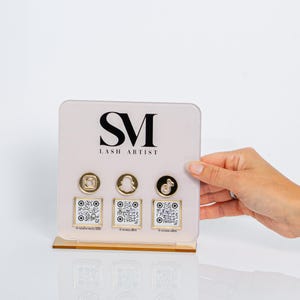 Peut inclure: Un présentoir en acrylique transparent avec une base dorée. Le présentoir présente les lettres "SM" au-dessus de "LASH ARTIST" et trois icônes de médias sociaux avec des codes QR correspondants. Le présentoir est tenu par une main.